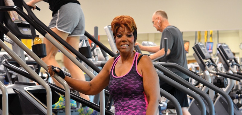 A smiling Black woman with orange hair wearing a purple and black tank top standing next to a Stairmaster