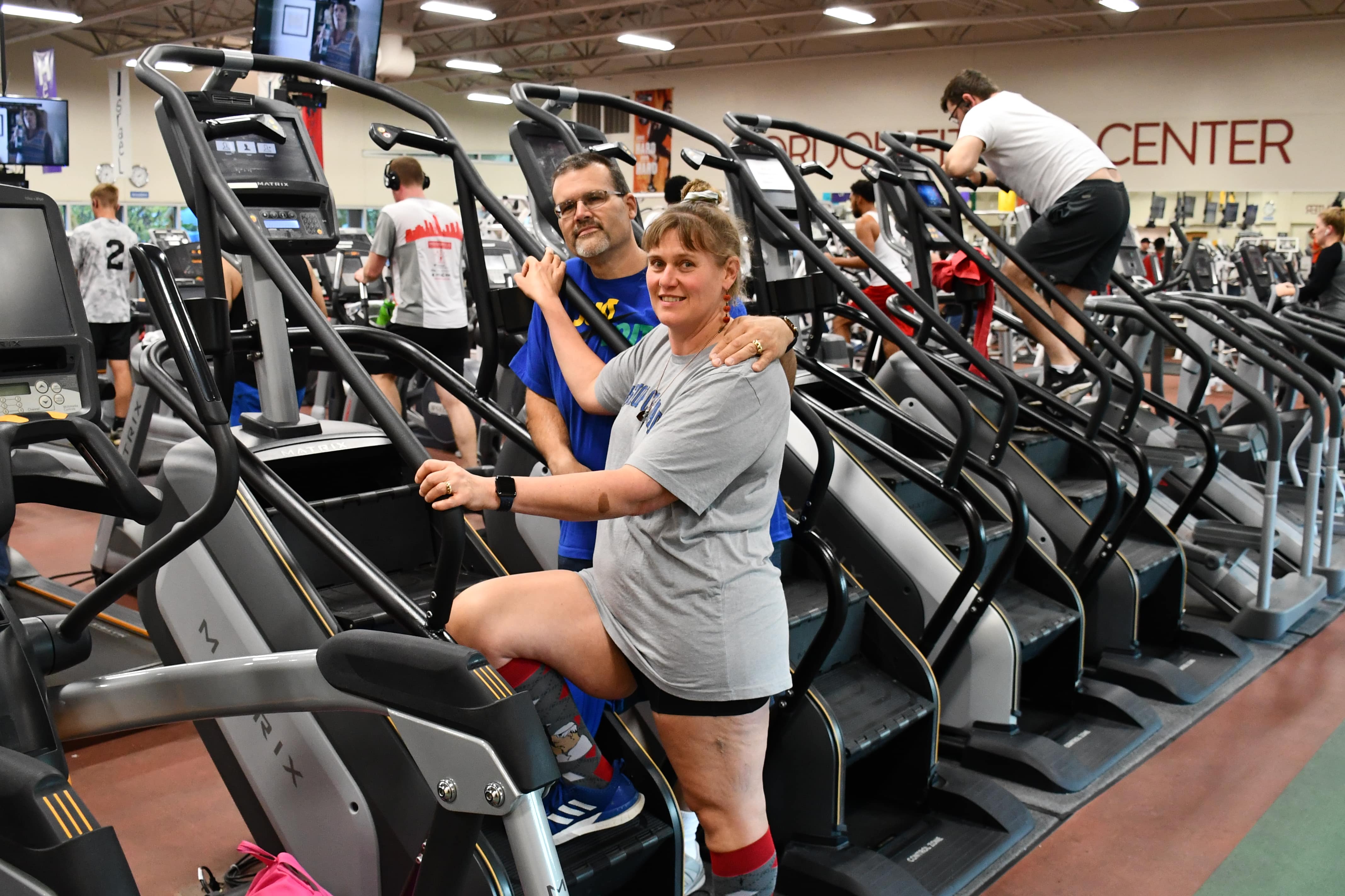 Andy and Laura Gzibovskis standing on a Stairmaster, with other JCC members working out in the background