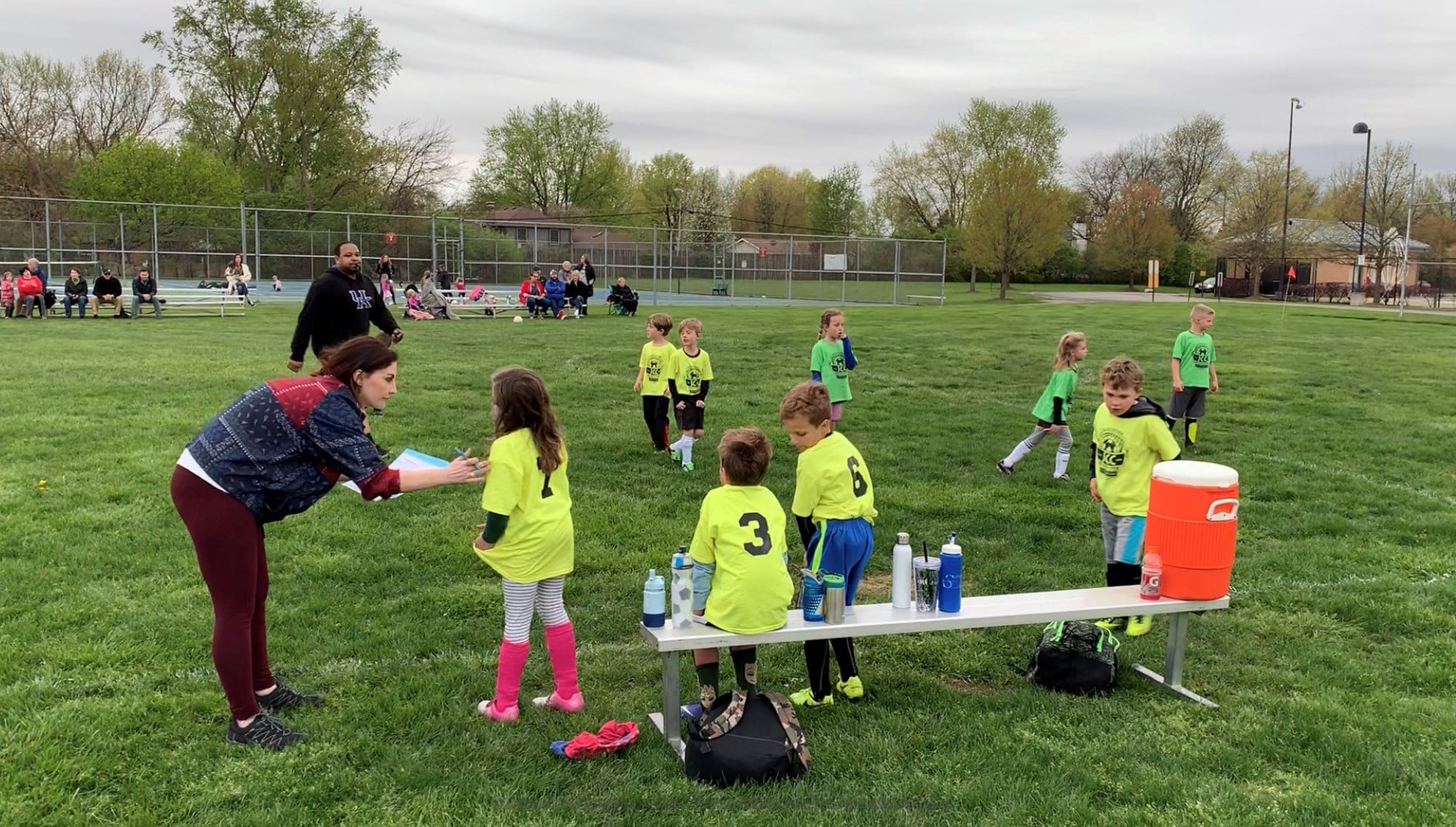 Jennie Carr (white woman in patterned sweater and maroon leggings) talking to a young girl in a yellow JCC soccer shirt, striped leggings and bright pink shin pads and cleats, as other children in yellow and green JCC soccer shirts take a water break.