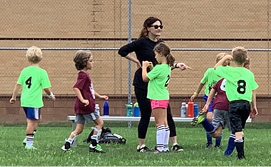 Jennie Carr (white brunette woman wearing all black) overseeing a group of children wearing green and maroon JCC soccer shirts