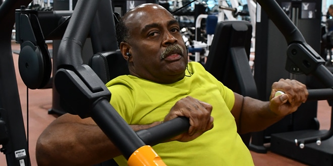 Joe Johnson (a mustached Black man in his 60s wearing a yellow shirt) working out on a machine in the Mordoh Fitness Center