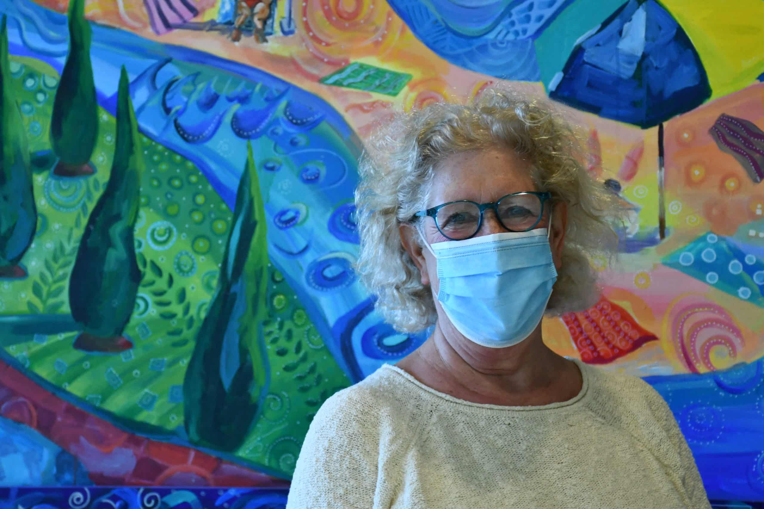 Cathy Kravitz (Jewish white woman with blonde hair in her 60s wearing an ivory sweater and mask) standing in front of a colorful mural