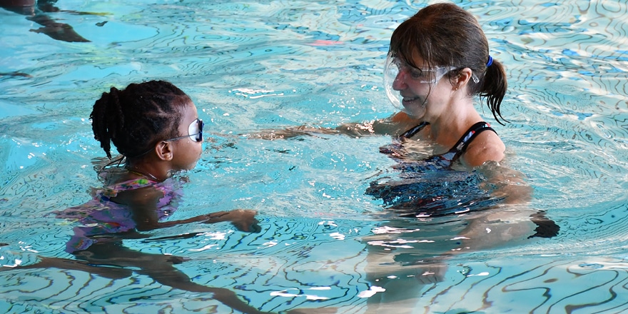 An older white woman with a ponytail of brown hair instructs a young black girl wearing goggles in the Backer Therapy Pool
