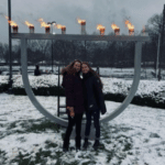 Two young white women in front of a giant, fully lit menorah on snow-covered ground on a cloudy winter day