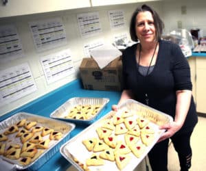 A gray-haired woman holding trays of hamantaschen