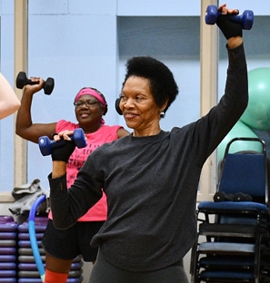 A Black woman in a long-sleeved gray shirt lifting dumbbells in a HIIT group ex class