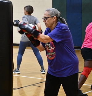 A grey-haired woman in a purple shirt hitting a punching bag in Total Body BoxJUMP group ex class.
