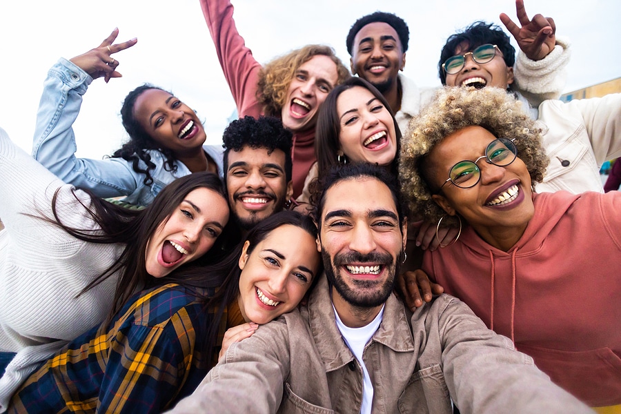 Group of friends taking a selfie outdoors on a sunny day, with clear skies in the background. The group is dressed in casual clothing, including jackets and hoodies, and they are all smiling and making peace signs.