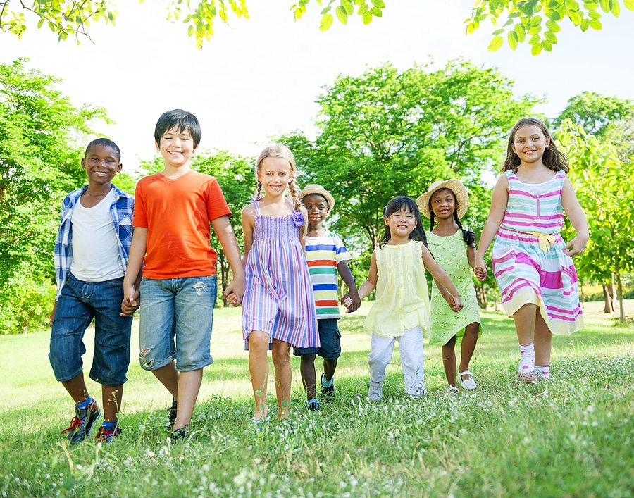 Seven elementary age children stand in a line, holding hands in a beautiful open outdoor area with green trees and grass.